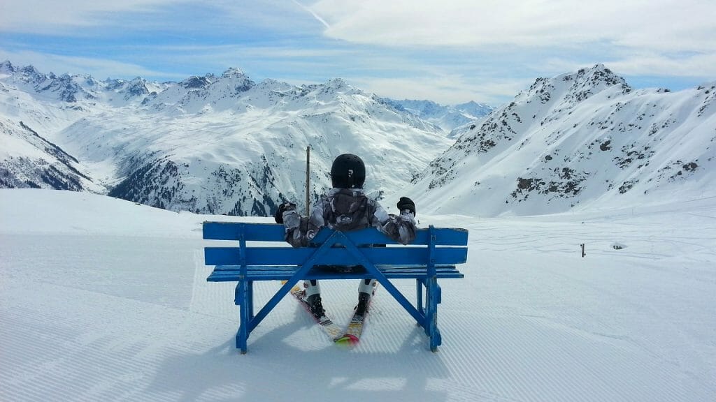 Person sitting with skis on blue bench in front of snowy ski slope and mountains in the distance