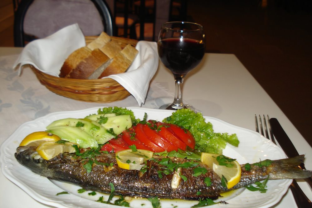 Whole fish with fresh tomatoes and cucumbers on a plate with a breadbasket and wine glass in the background