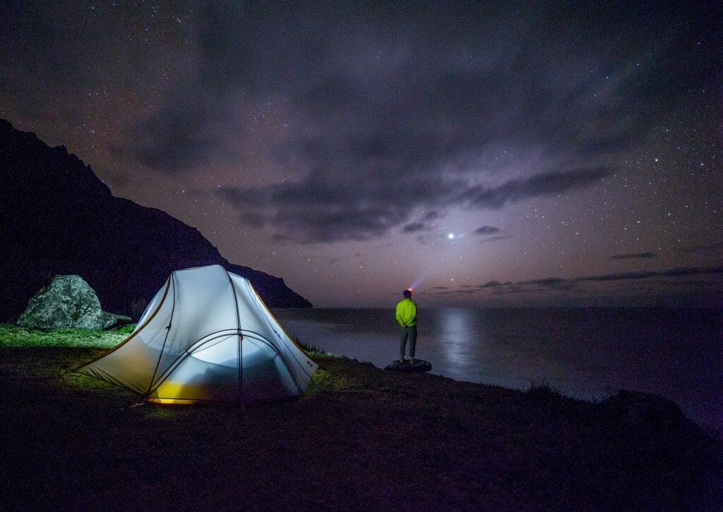 man with headlamp standing by a lake with tent next to him at night