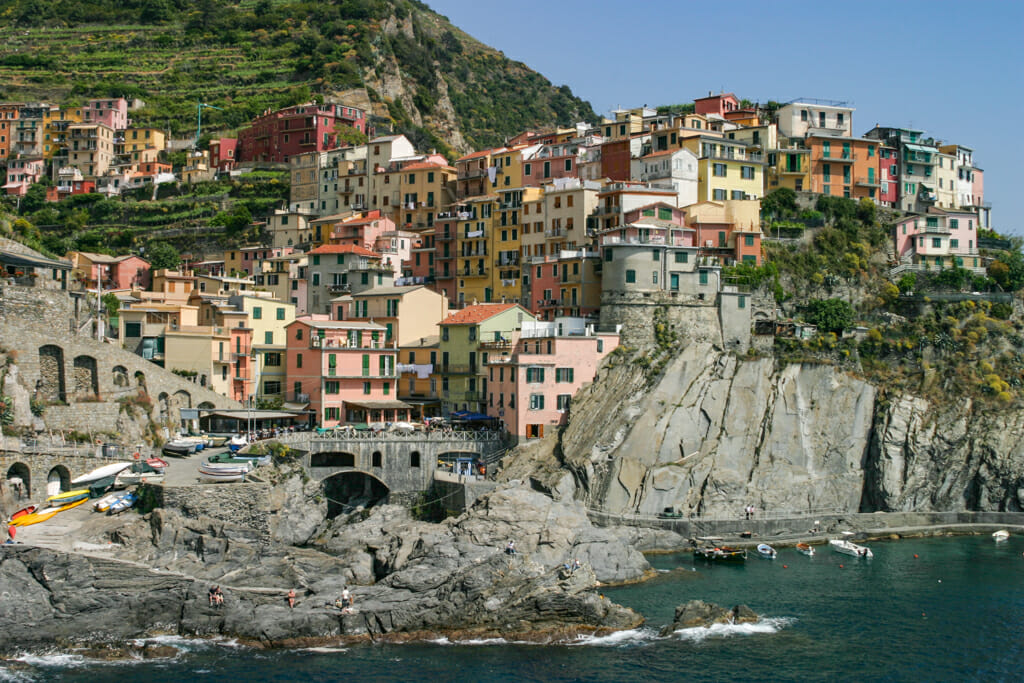 Cinque Terre Trail - A breathtaking view of Manarola perched on the cliffs depicting a lifestyle with colorful buildings, swimmers, and boats in the cove.