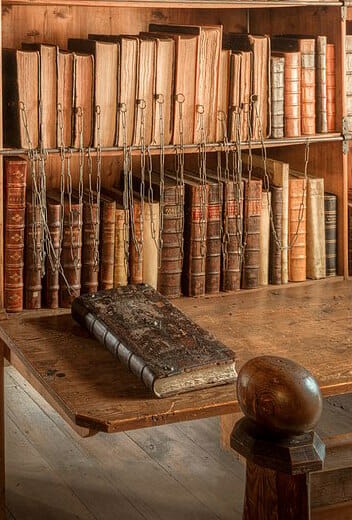 A book shelf of chained books with one pulled out, laying on the desk to read