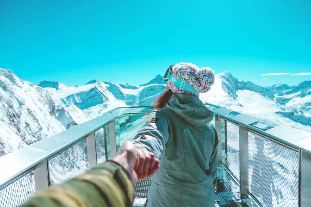 Woman in winter clothes reaching back for mans hand in front of snow capped mountains