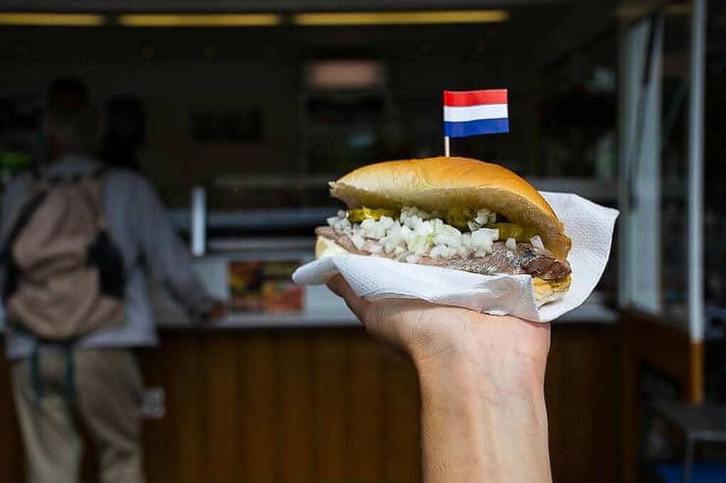 Man holding a herring sandwich with onions and topped with the flag of Holland