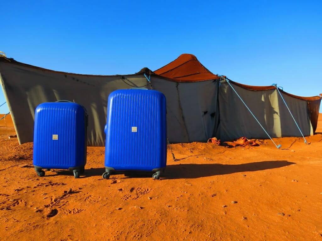 Two bright blue hard shell suitcases stark against the orange sand in front of a gray tent