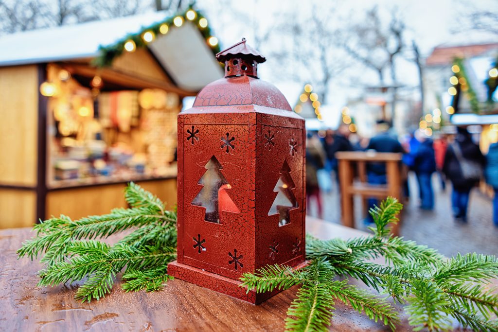 Lantern on a table surrounded by pine branches with the lively christmas market in the background