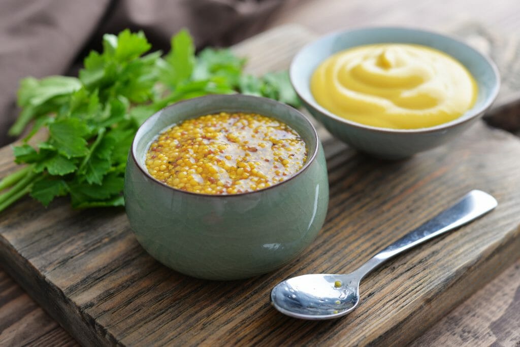 2 Varieties of Dijon Mustard in little bowls on a wood board with spoon and herbs in the background