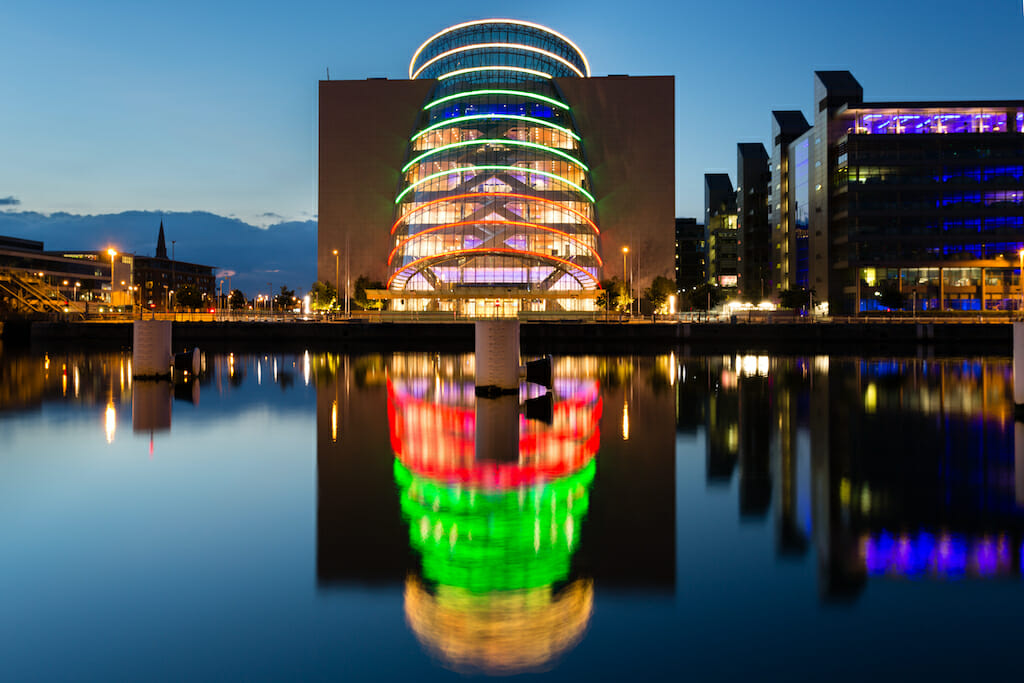 The bright lights on the convention centre in Dublin shine brightly and reflect in the water below, the lights from the other buildings aren't as bright in the early night