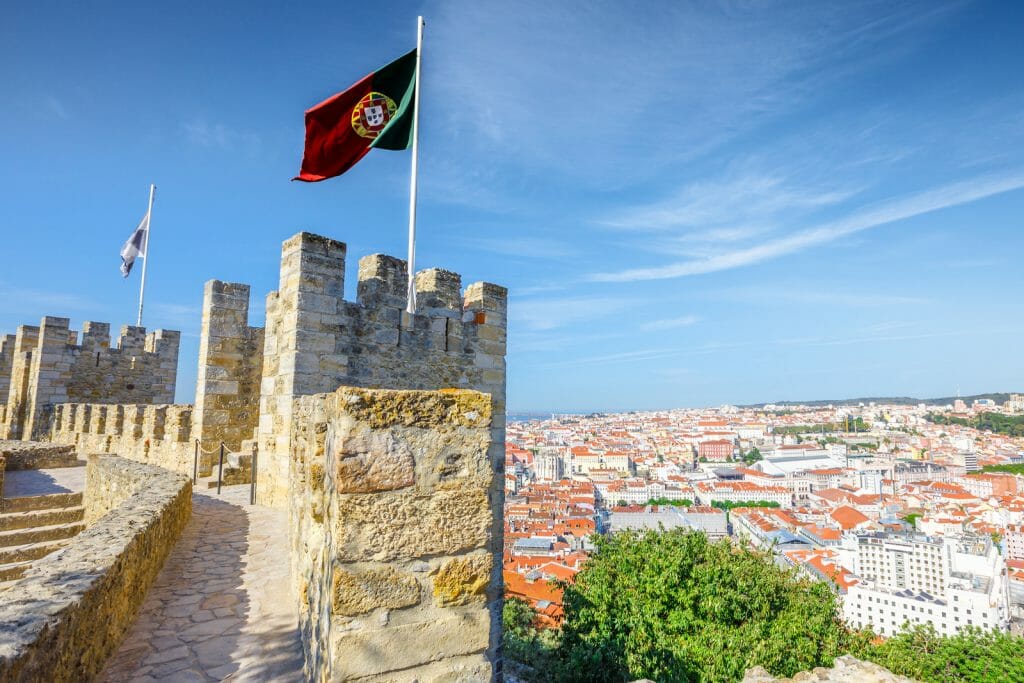 Portugal flag waving on ancient fortress wall of Sao Jorge Castle, at Moorish castle on highest hill in Alfama. Lisbon aerial cityscape on background. Lisbon Castle is a popular tourist attraction.
