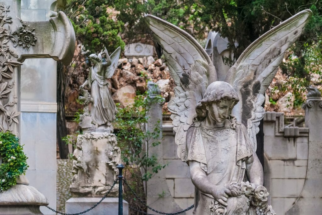 The sculpture of angel on the background of graves and another sculpture on the Montjuic Cemetery, Barcelona, Catalonia, Spain