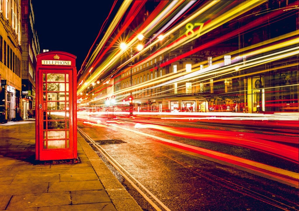 Long exposure photo showing red phone booth in London and light rails of a red double-decker bus passing by on the right