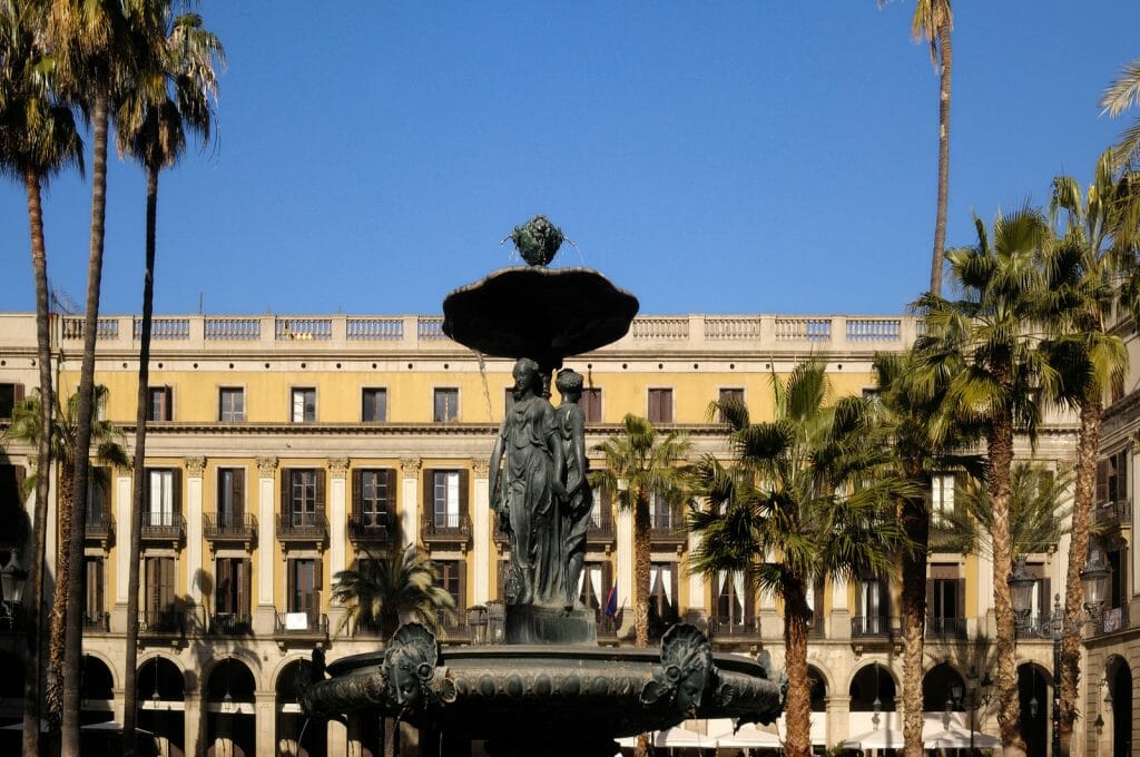 Plaça Reial, metal fountain in front of large yellow building with many columns in the background, square with palm trees, Barcelona, Spain
