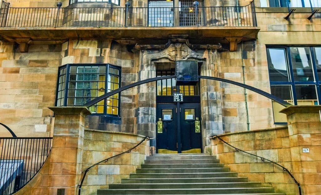 A building designed by Charles Rennie Mackintosh in Glasgow. Yellow and orange sandstone building with a narrowing staircase leading to a decorative black door. 