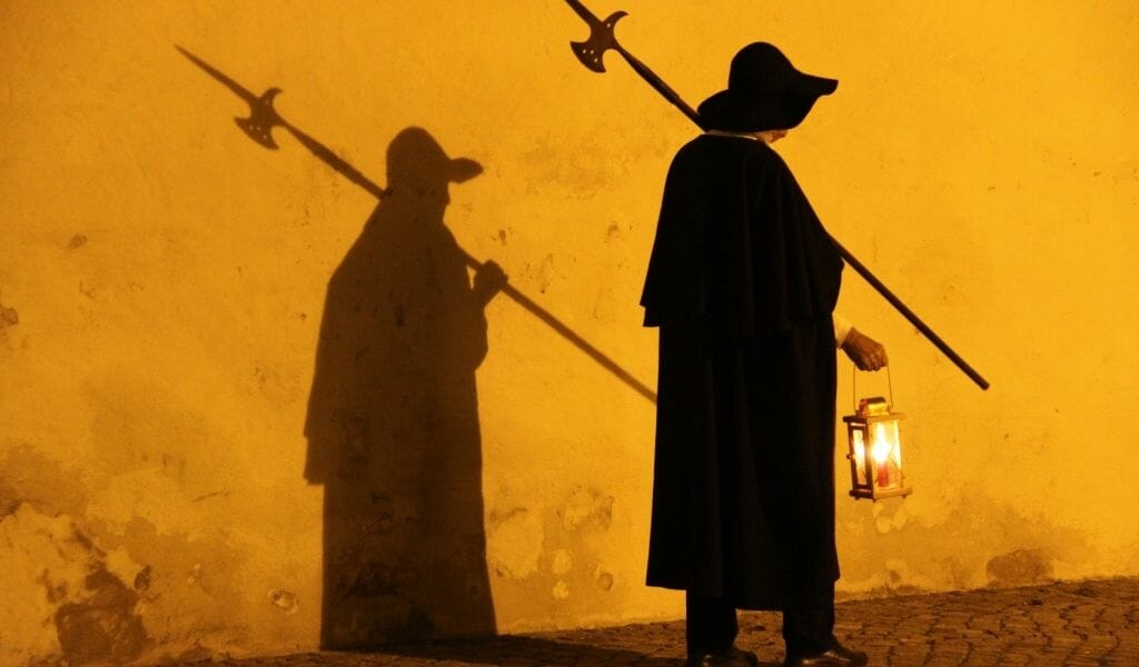 Nightwatchman in a coak and with a spear and lanter casting a shadow on an orange wall in Rothenburg ob der Tauber