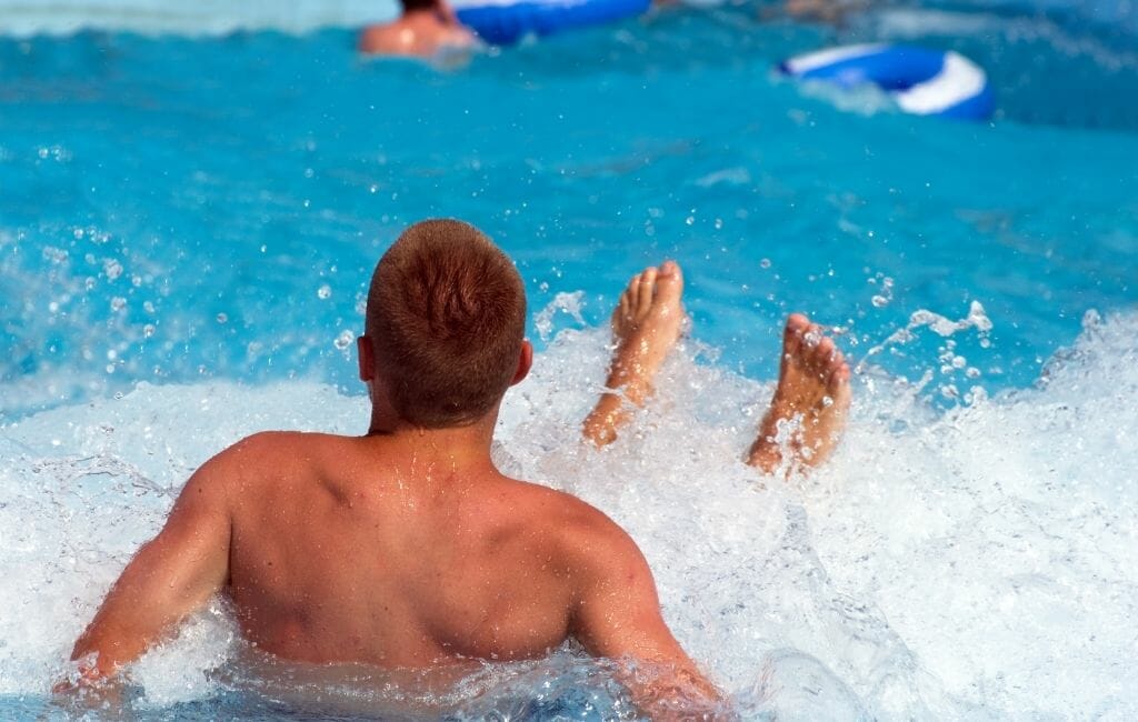 Man sitting with his back to camera in a wave pool with a wave crashing into him