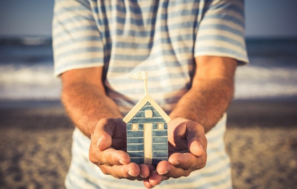 Closeup of man holding a small ceramic house in his hands with beach in the background