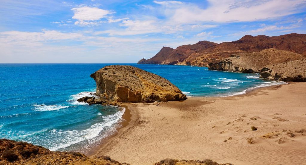 Large rock formations coming out of otherwise flat, sandy beach next to bright blue waters of the Mediterranean sea with desert mountains in back on a sunny day. Playa del Monsul Beach, Cabo de Gata, Almeria, Spain. Best beaches in Almeria.