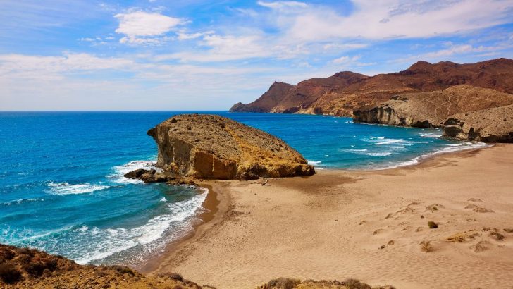Large rock formations coming out of otherwise flat, sandy beach next to bright blue waters of the Mediterranean sea with desert mountains in back on a sunny day. Playa del Monsul Beach, Cabo de Gata, Almeria, Spain. Best beaches in Almeria.