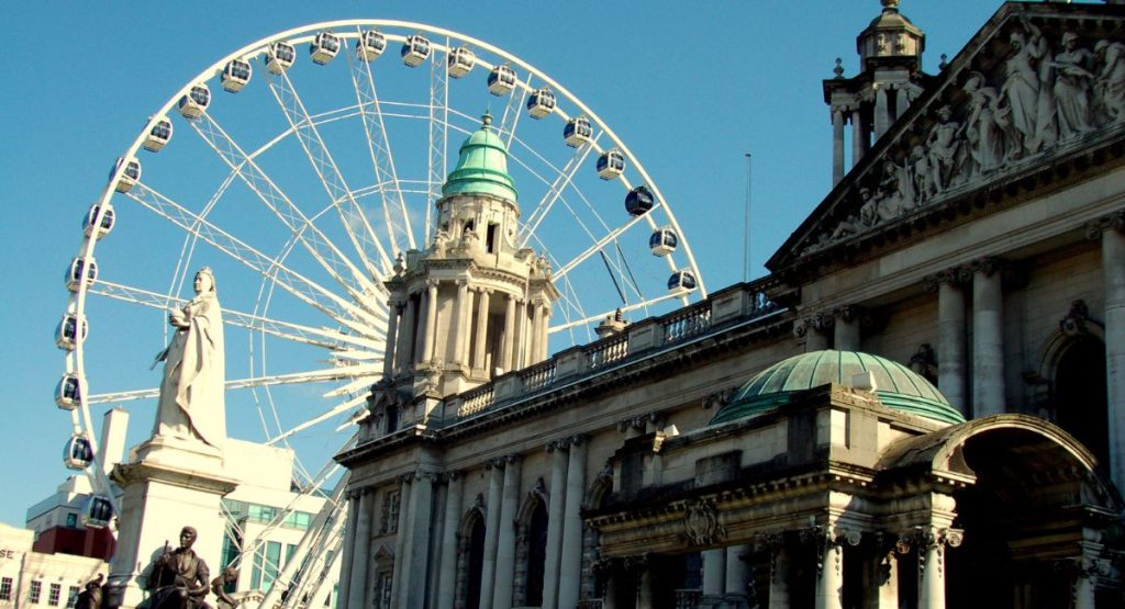 Large ferris wheel/observation wheel rising up behind historic building and statues of Baroque Revival style. Belfast City Hall and Belfast Wheel, Belfast, Northern Ireland.