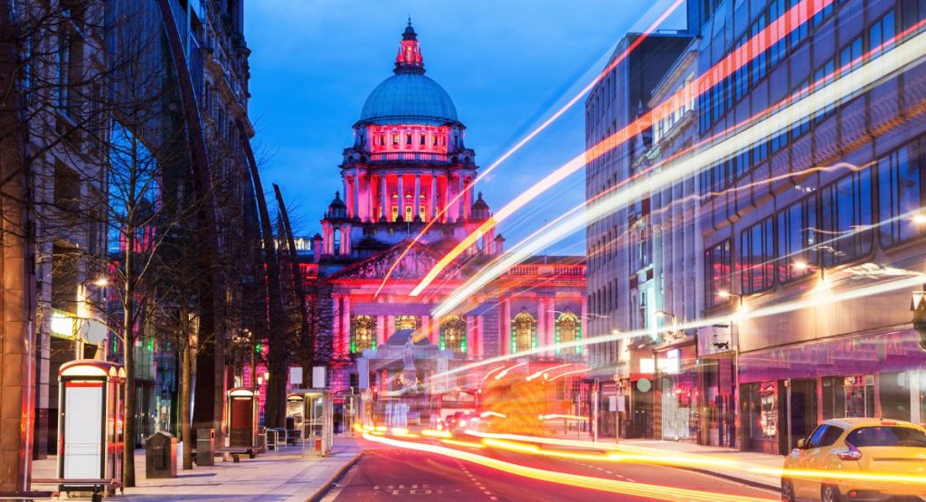 Night shot of busy street in downtown Belfast with multi-story buildings on either side of roadway and historic domed-building, Belfast City Hall, at end of road. Belfast, Northern Ireland.