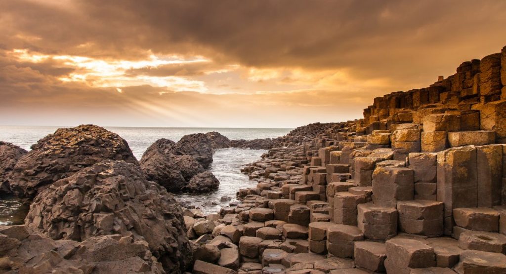 Grey basalt columns stacked like stairs along shoreline of Atlantic Ocean during orange sunset. Giant's Causeway, Bushmills, Northern Ireland.