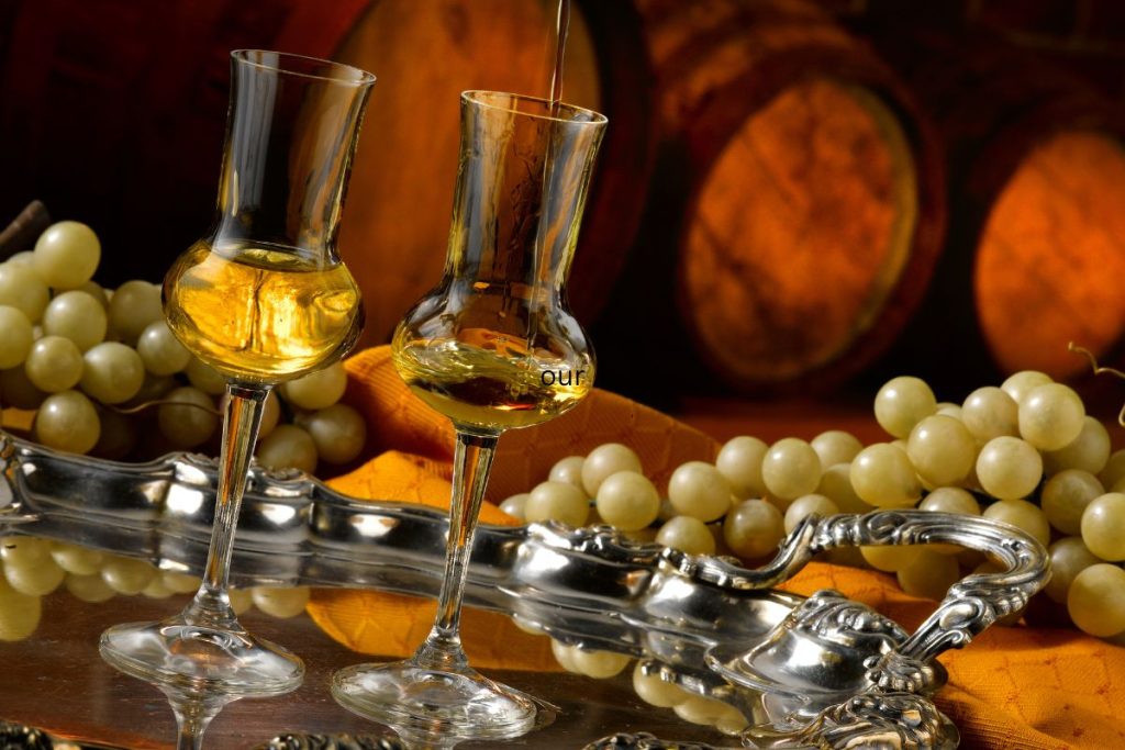 two glasses of italian grappa on a silver tray with white wine grapes and barrels in the background