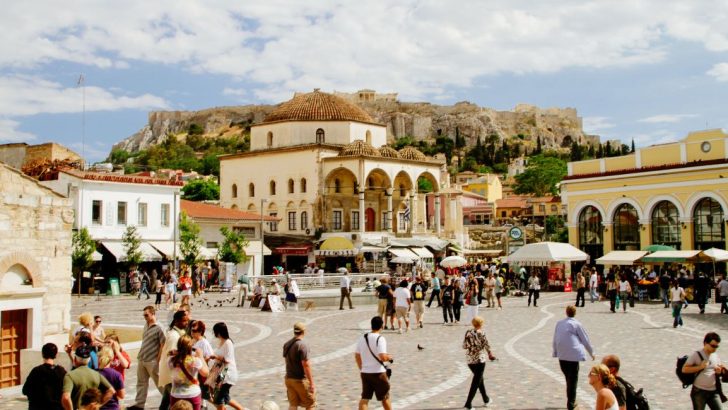 Courtyard square surrounded by low rise buildings with shopping and restaurants, square is filled with pedestrians on sunny summer day, Greek ruins stand atop hill in background. Monastiraki Square, Athens, Greece, What to do in Athens in 3 Days.