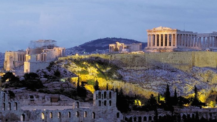Huge, white Greek ruins on hilltop overlooking city. Ruins are white and have large, intricate pillars and arched windows at nighttime. Parthenon in Athens, Greece. 3 Days in Athens.