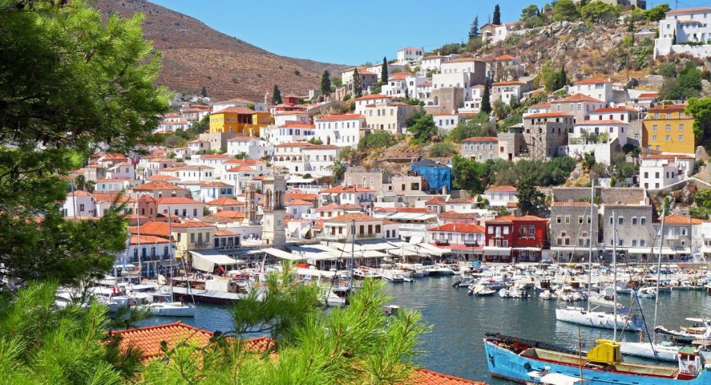Hilltop filled with white buildings with orange roofs overlooking crowded harbor full of boats including fishing boats and sailboats on sunny summer day. Port Town, Hydra Island, Greece, Greek Islands.