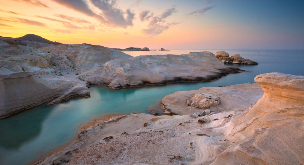 White smooth rock shoreline over turquoise, calm water of Aegean Sea during colorful sunset. Sarakiniko Beach, Milos Island, Greece. Best islands in Greece Islands.