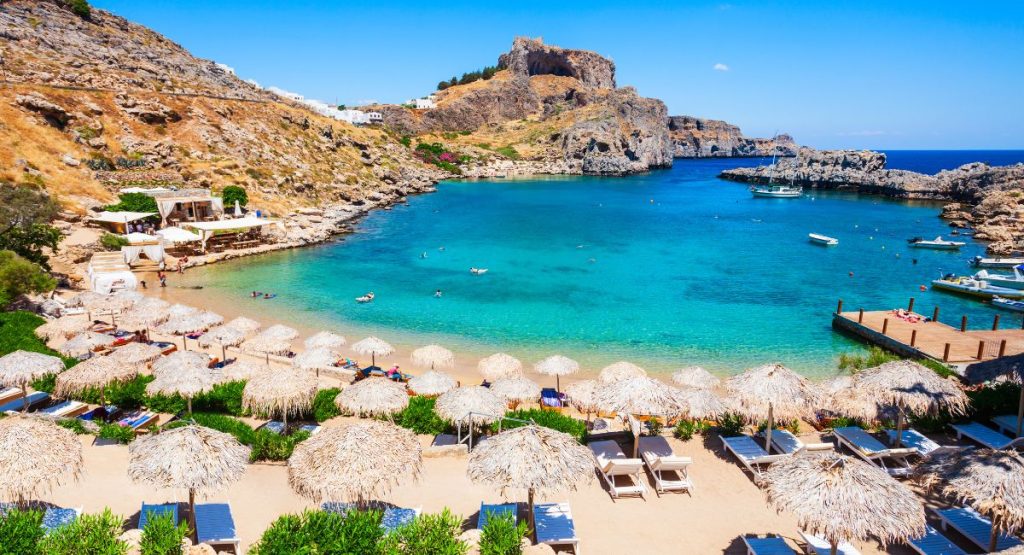 Clear blue water in cove surrounded by rocky shoreline with strip of sandy beach, beach is covered in straw umbrellas and beach chairs on sunny summer day. Rhodes Island, Greece, Best Greek Islands.