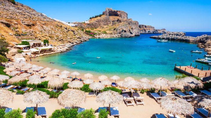 Clear blue water in cove surrounded by rocky shoreline with strip of sandy beach, beach is covered in straw umbrellas and beach chairs on sunny summer day. Rhodes Island, Greece, Best Greek Islands.
