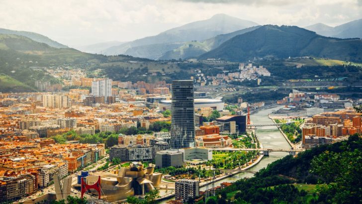 City center alongside river with green mountains in background. Where to stay in Bilbao Spain.