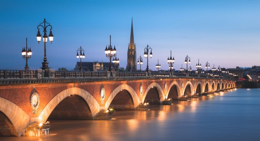 Arched bridge stretching across wide, flat river during sunset with tall steepled church in background. Where to stay in Bordeaux, France.