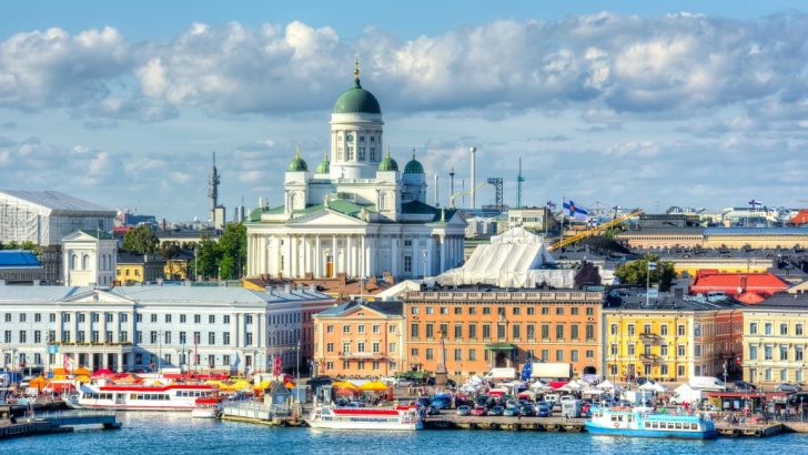 City skyline with colorful buildings, large white and green domed cathedral sitting on waterfront with boats on sunny day. Where to stay in Helsinki, Finland.